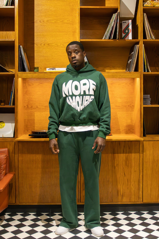 Person wearing a green tracksuit with 'MORE AMOURE' text in a room with wooden shelves and a checkered floor.