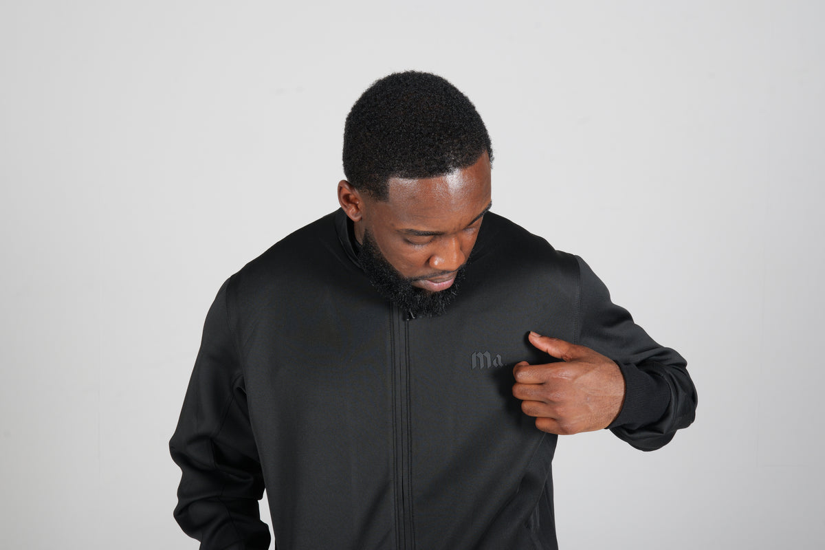 Close-up of a man in the black More Amoure Core Zip Jacket touching the small tonal logo on the chest