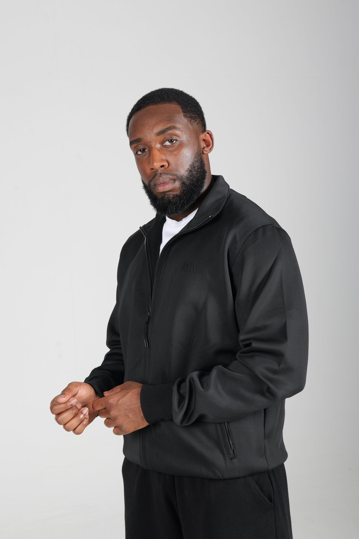 Man in the black More Amoure Core Zip Jacket with a white tee underneath, hands together, in a relaxed studio pose