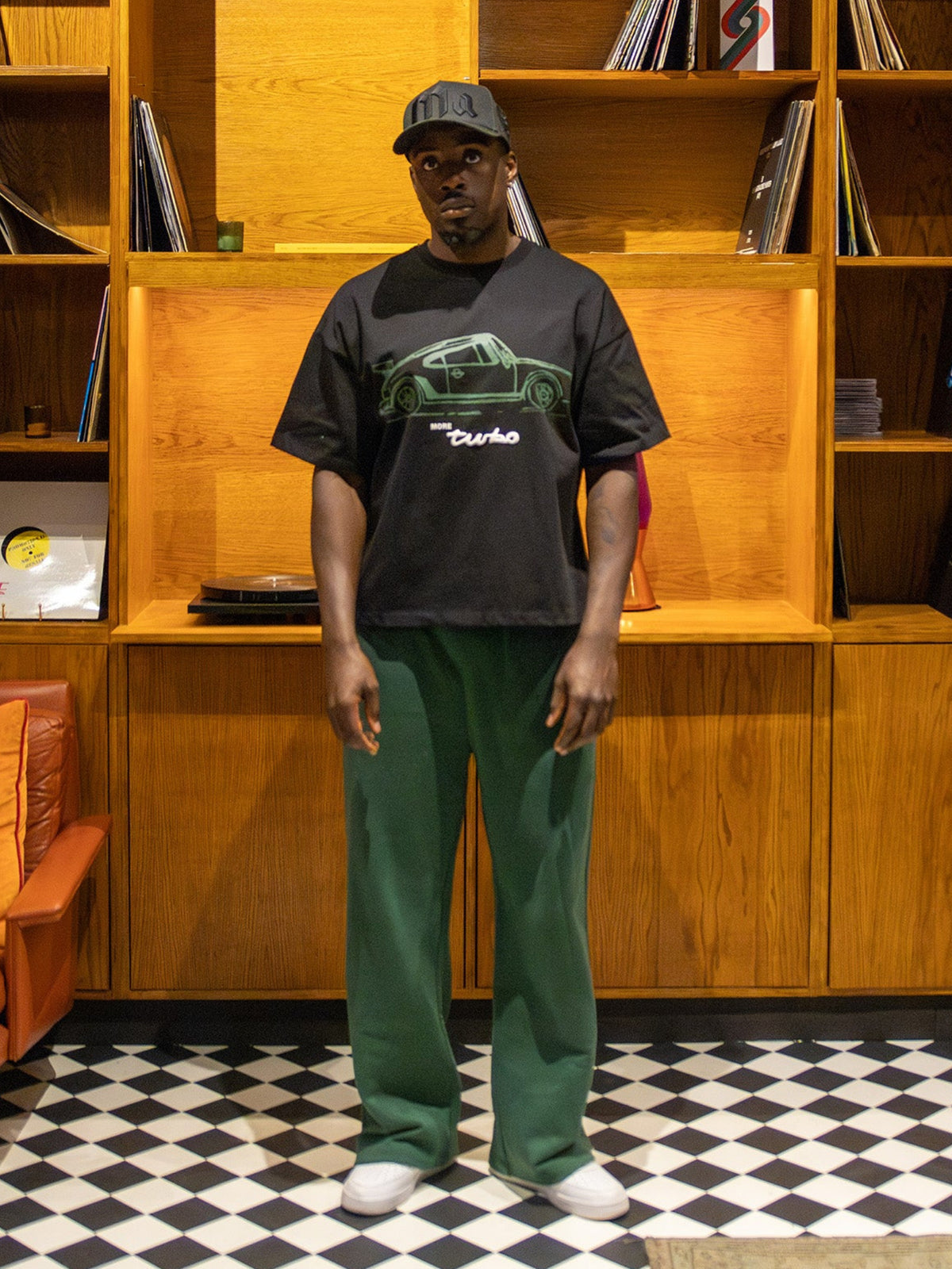 Person wearing a black t-shirt with a graphic design and green pants standing in front of a wooden bookshelf.
