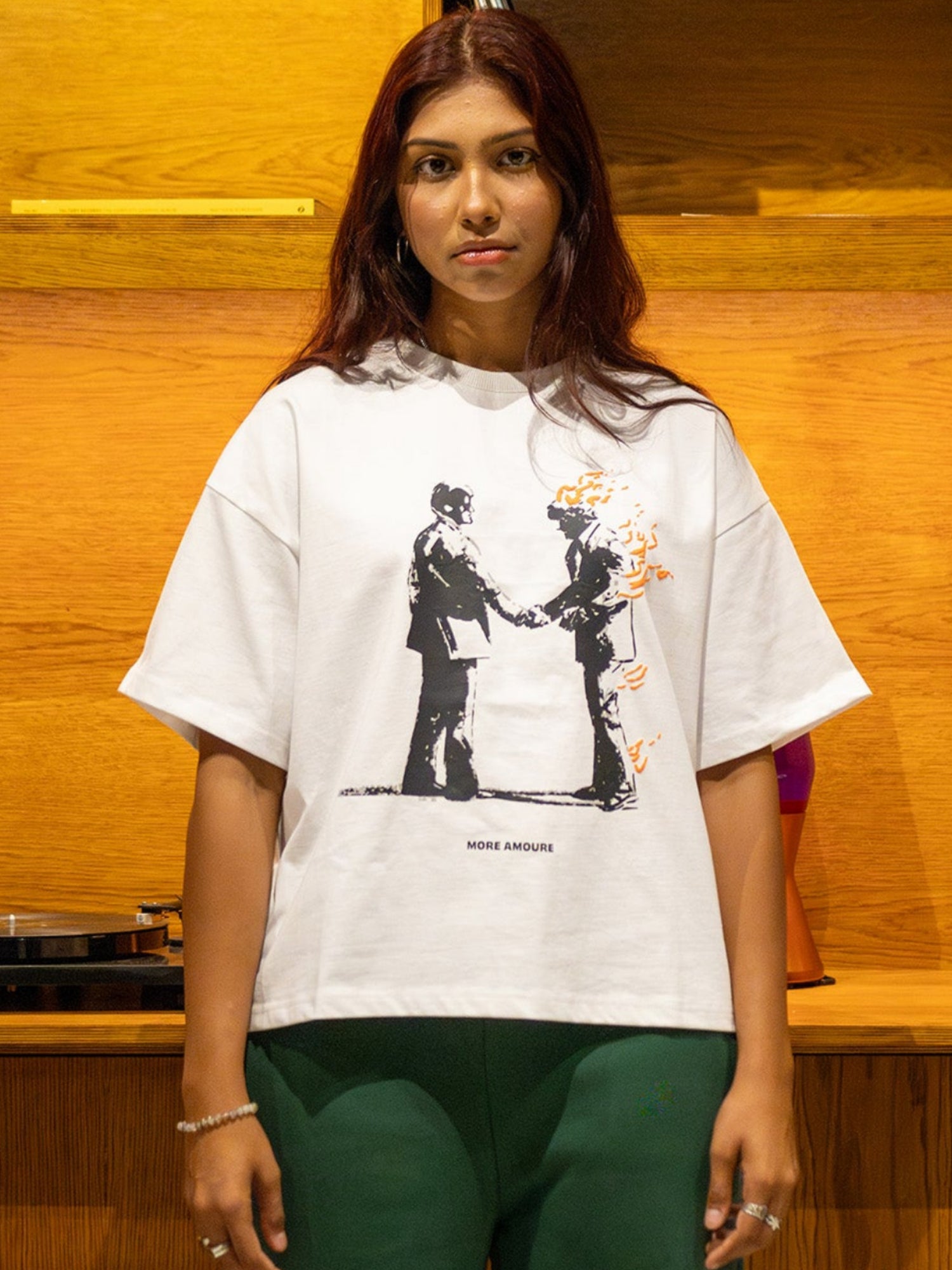 Person wearing a white t-shirt with graphic design in front of a wooden shelf.