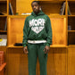 Person wearing a green tracksuit with 'MORE AMOURE' text in a room with wooden shelves and a checkered floor.