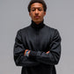A man wearing a black zip-up jacket with a high collar, standing with arms crossed against a solid gray studio background.