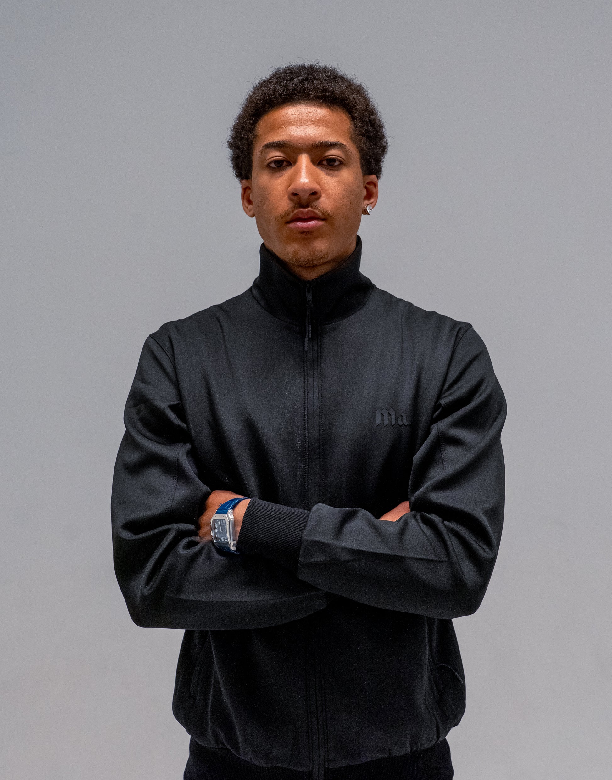 A man wearing a black zip-up jacket with a high collar, standing with arms crossed against a solid gray studio background.