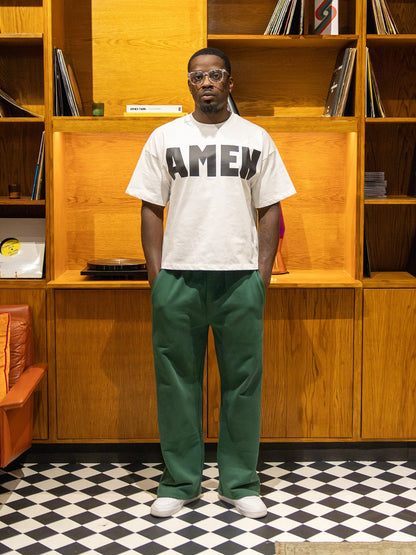 Person wearing a white t-shirt with 'AMEN' text and green pants standing in front of a wooden bookshelf.